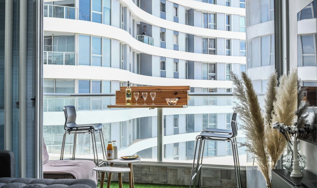 Un interior modern amb un balcó corbat, una taula de fusta amb cadires, i vista a un edifici alt.