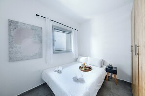 A modern hotel room with a bed, a side table, a tray with toiletries, a window with curtains, and a wall art piece.