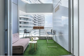 A balcony with a green artificial turf floor, a sofa, two chairs, and a small table with a bottle and glasses.