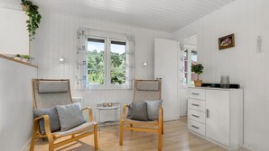 Une pièce comprenant deux chaises en bois avec des coussins, une petite table ronde, un meuble blanc avec des tiroirs et une fenêtre avec des rideaux.