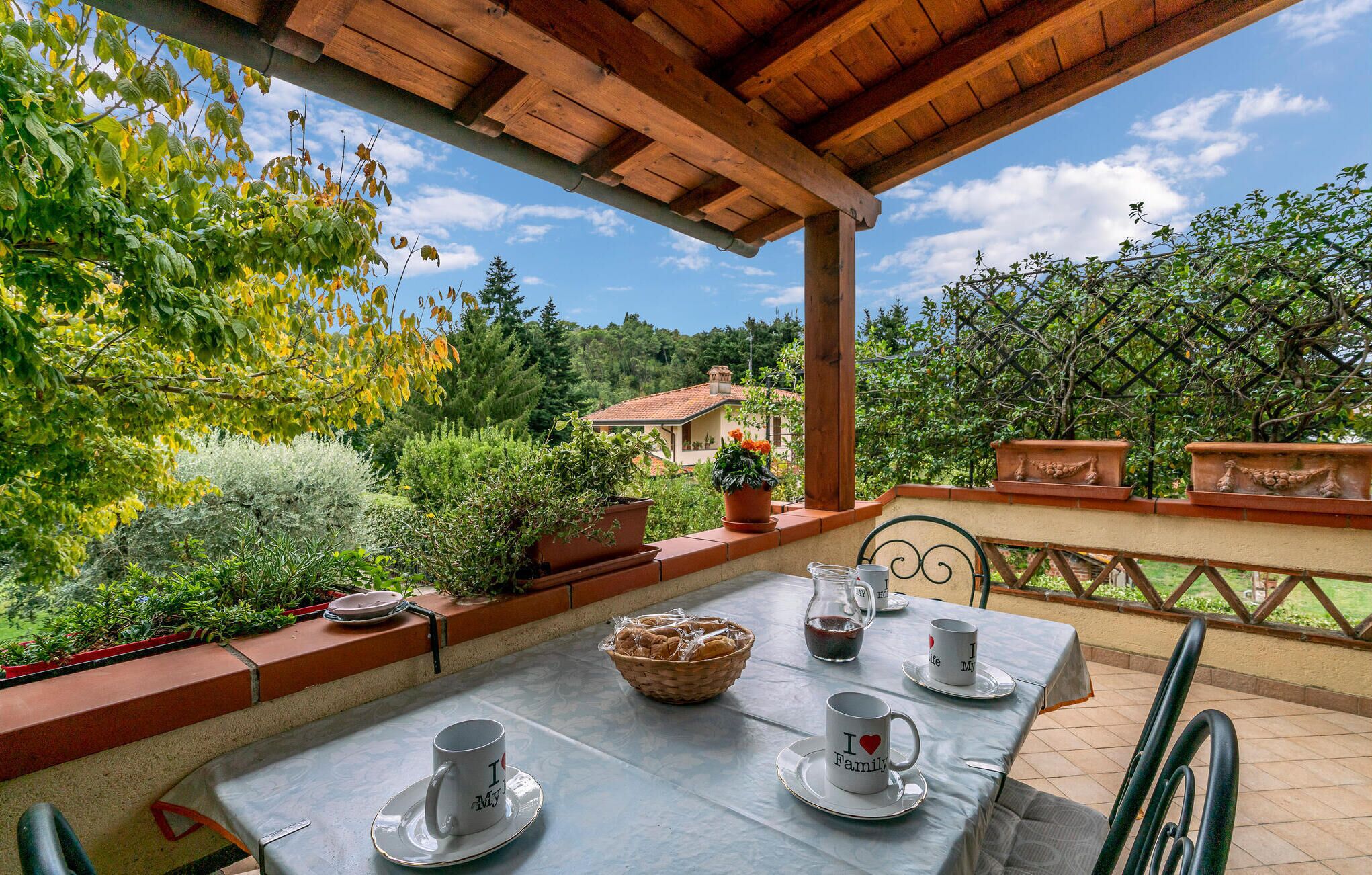 An outdoor patio with a table set for two, surrounded by potted plants and a scenic view of a house and trees.