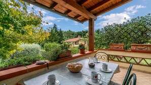 Eine Terrasse im Freien mit Tisch für zwei Personen, umgeben von Pflanzen in Töpfen und einem schönen Blick auf ein Haus und Bäume.