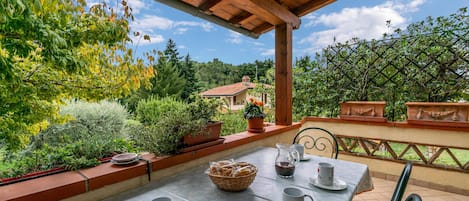 Eine Terrasse im Freien mit Tisch für zwei Personen, umgeben von Pflanzen in Töpfen und einem schönen Blick auf ein Haus und Bäume.
