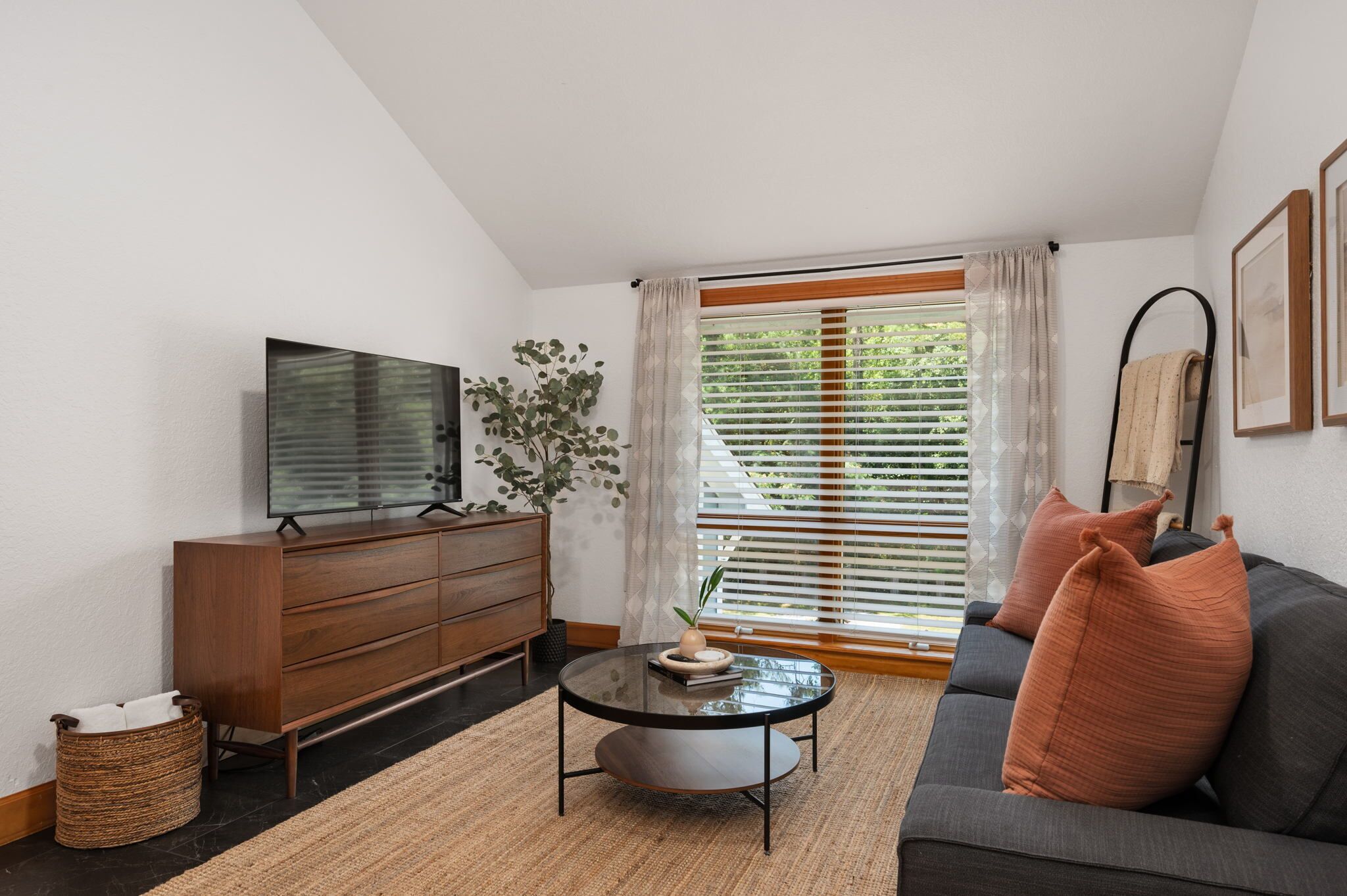 A modern living room with a flat-screen TV, a wooden cabinet, a round coffee table, a sofa with cushions, and a large window with blinds.
