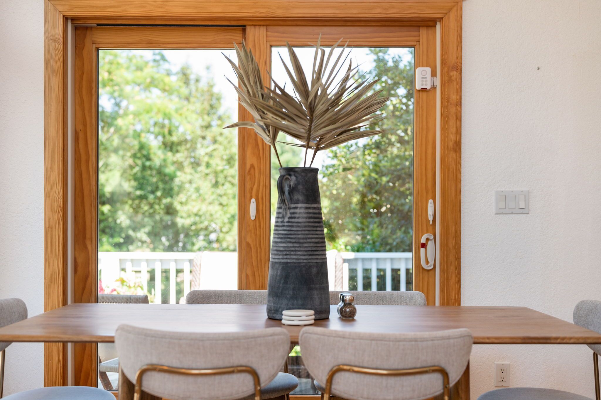 Een eethoek met een houten tafel en twee stoelen, een vaas met gedroogde planten, en uitzicht op groen door grote ramen.