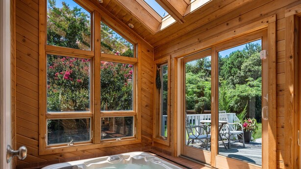 A wooden cabin with a hot tub, large windows, and a deck with outdoor furniture. - Indoor Hot Tub, Barrel Sauna, Pool, Natural Light! (Powells Point)
