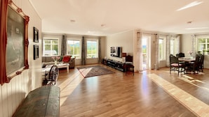 A spacious living room with wooden flooring, a sofa, a coffee table, and a TV. There are large windows with curtains, a dining area with chairs, and a view of greenery outside. - Amazing home in Lunde with WiFi (Lunde)