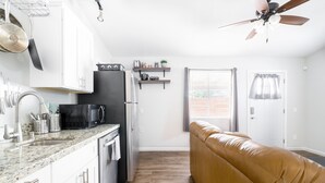 Ett modernt kök med vita skåp, ett svart kylskåp och en bänkskiva i marmor. I vardagsrummet finns en brun lädersoffa och en takfläkt.