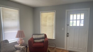 Un salon avec un canapé à rayures, un fauteuil rouge, une lampe et un tapis. Il y a deux fenêtres avec des stores et une porte blanche.