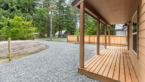 Une terrasse en bois couverte, avec une allée de gravier menant à une maison entourée d’une clôture en bois et d’arbres denses.