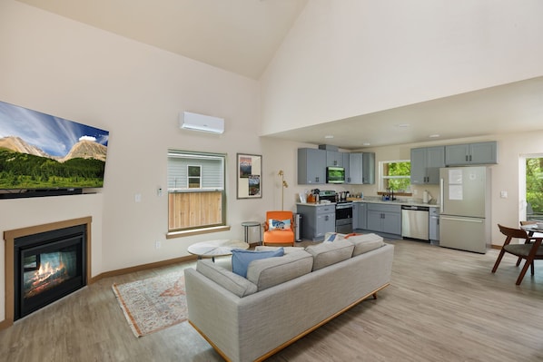 A spacious living area with a fireplace, a sofa, a dining table, and a kitchen with grey cabinets and a refrigerator.