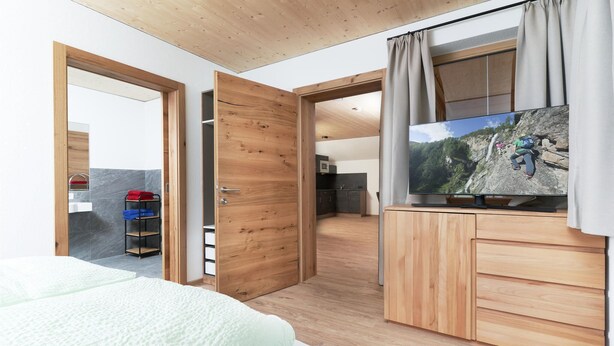 Een moderne slaapkamer met een houten plafond, een groot bed, een televisie op een houten dressoir en uitzicht op een keuken en terras.