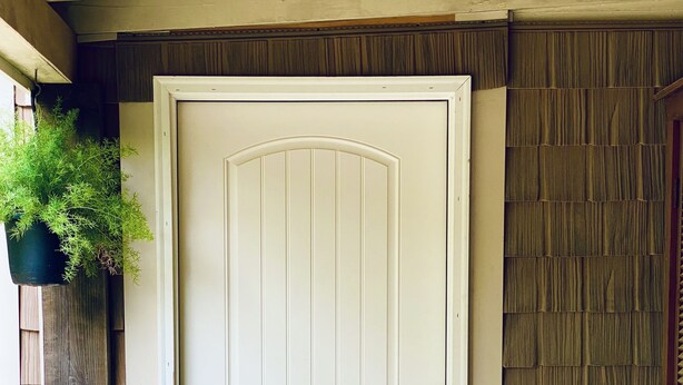 Une porte d’entrée blanche avec une petite fenêtre, un paillasson sur lequel est écrit « BONJOUR », et une clôture en bois sur la gauche.