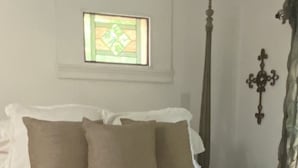 Un lit avec une literie blanche et beige, une petite fenêtre ornée de vitraux, un long poteau en bois et un élément décoratif fixé au mur.
