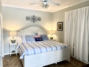 Un dormitorio con una cama blanca, ropa de cama a rayas, dos lámparas de noche, un ventilador de techo y un adorno en la pared.