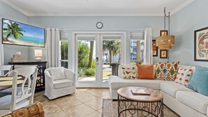 A living room with a white sofa, a coffee table, a TV, and a dining area with a table and chairs. - Shore Beats Work condo @ Caribbean Dunes across from Crystal Beach - Snowbirds ❤️ (Destin)