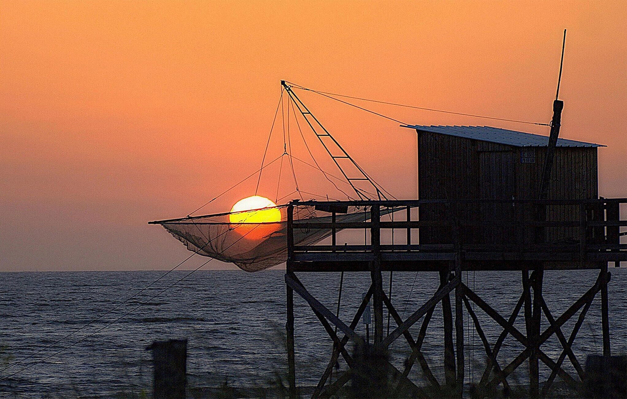 A wooden structure with a fishing net over the water, a wooden building, and a sunset in the background.