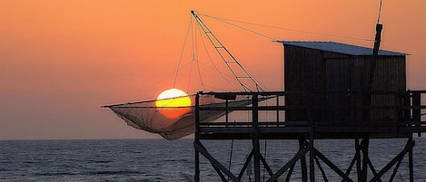 A wooden structure with a fishing net over the water, a wooden building, and a sunset in the background.