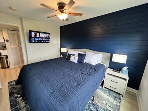 A bedroom with a dark blue bedspread, a ceiling fan, a TV mounted on the wall, and a bedside table with a lamp and tissues. - Stunning Views! Indoor Pool, Sleeps 2 (Gatlinburg)