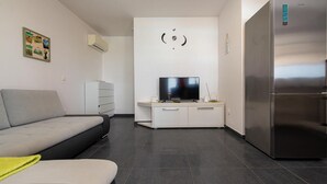 En moderne stue med en grå sofa, et fladskærms-tv på et hvidt skab, et rustfrit stål køleskab og en vægmonteret aircondition.