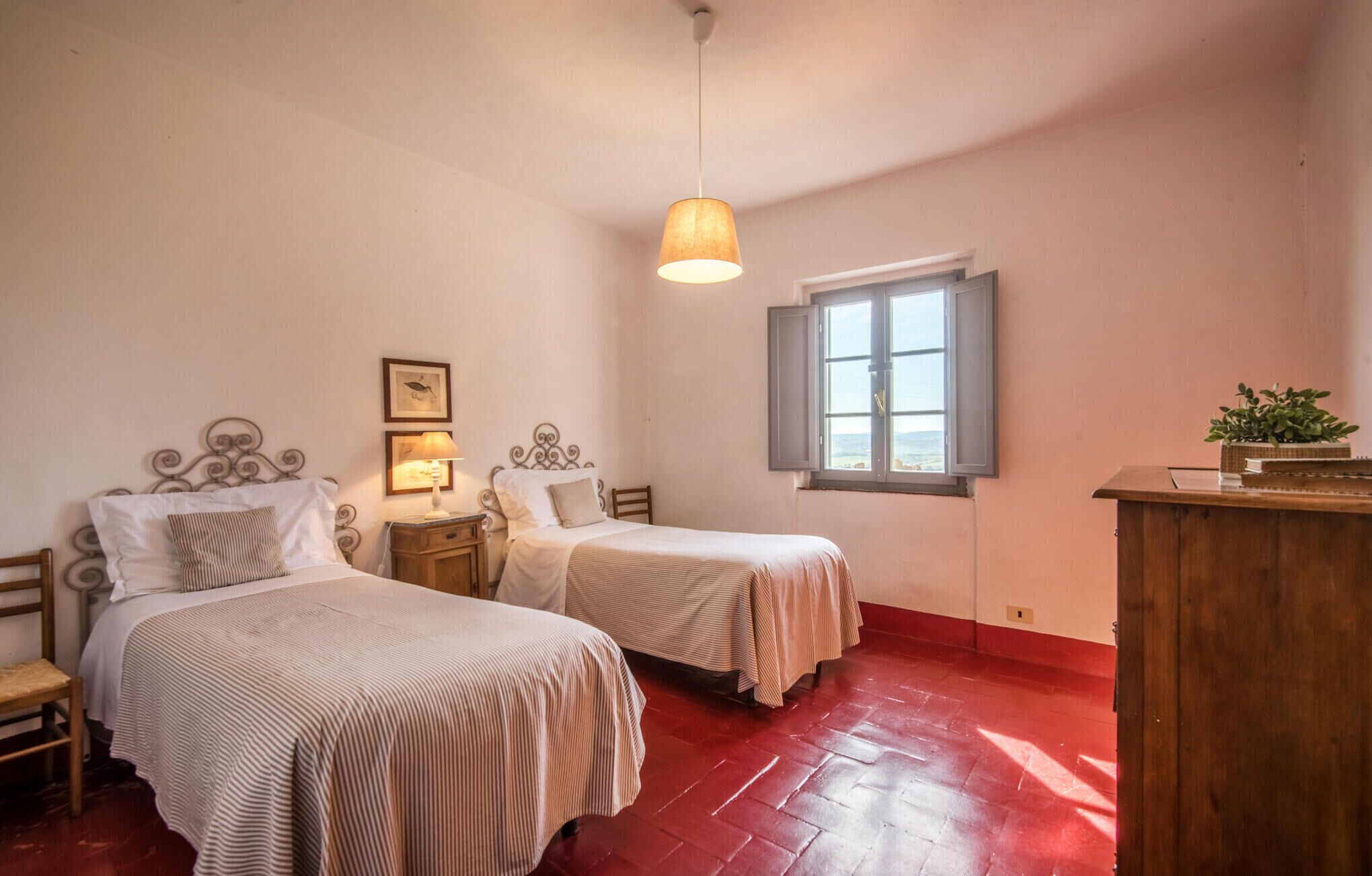 Quarto com duas camas, uma janela com persianas, um candeeiro pendente e uma secretária de madeira com uma planta.