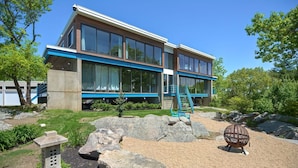 A modern two-story building with large windows, a blue exterior trim, and a landscaped garden with rocks and a small statue. - Secluded 20 acre 10BR ultra contemporary retreat (Manchester-by-the-Sea)