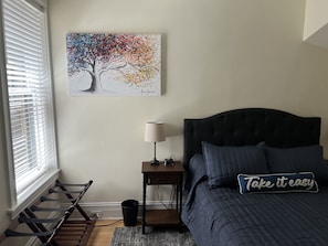 Una camera da letto con un letto, un comodino, una lampada, un quadro appeso al muro, una finestra con persiane e uno stendibiancheria.