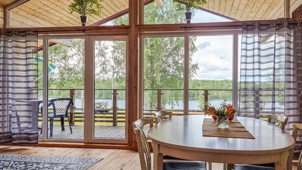 A wooden cabin with a large glass door leading to a deck overlooking a lake. Inside, there is a round table with chairs and a vase of flowers. - Awesome apartment in Färgelanda (Färgelanda)