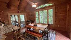 A rustic wooden cabin with a dining area featuring a large table and benches, a kitchen area with a marble countertop, and large windows offering a view of the surrounding forest. - Spacious cabin w/Hot Tub , 10 minutes from downtown Blue Ridge. (Cherry Log)