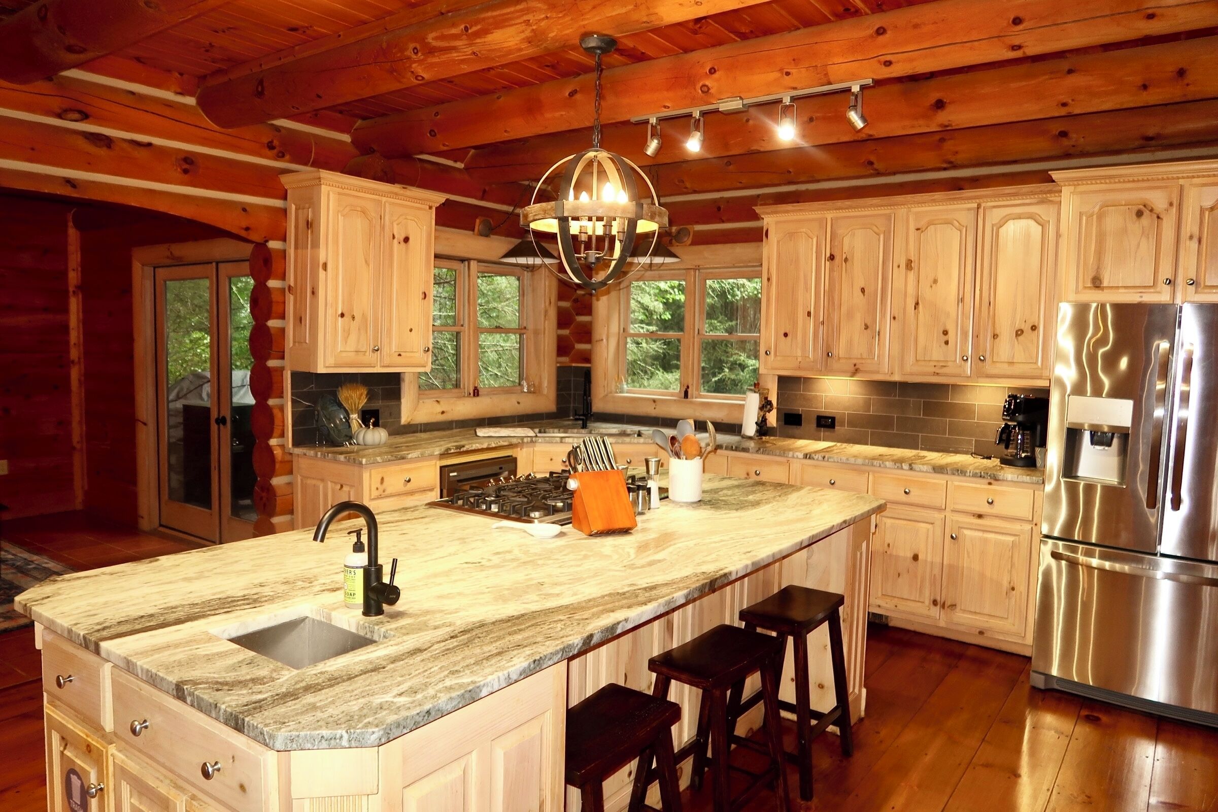 A spacious kitchen with wooden cabinets, a large island with bar stools, a stainless steel refrigerator, and a hanging light fixture.