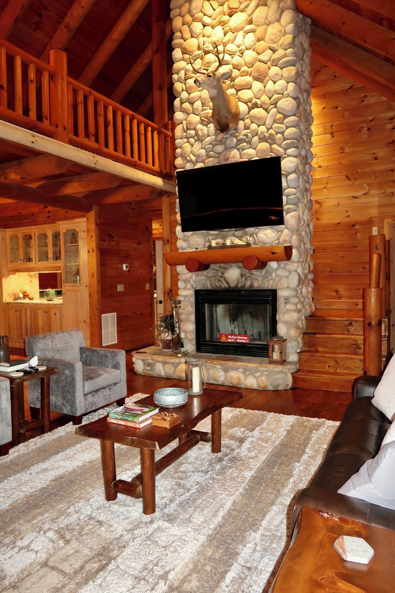 A cozy log cabin interior with a stone fireplace, wooden furniture, and a mounted deer head.