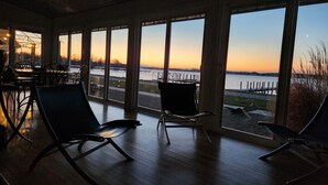Una habitación con ventanas amplias que dan a un atardecer y una masa de agua. Hay dos sillas y una mesa en la habitación.