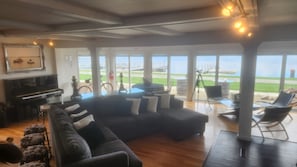 A spacious living room with a large sectional sofa, a piano, and a view of the beach through large windows.