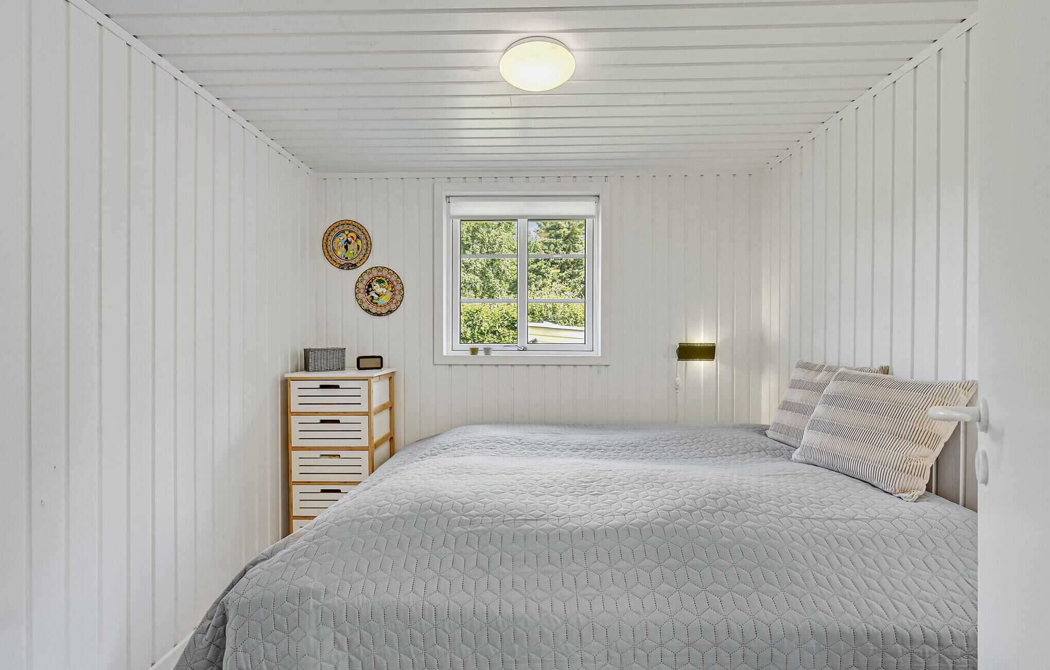 Een slaapkamer met een bed, nachtkastje, lamp en raam met uitzicht op groen.