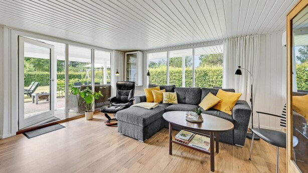 A spacious living room with a grey sofa, yellow cushions, a wooden coffee table, and a black chair. Large windows offer a view of a garden and a white ceiling with horizontal lines. - Cozy home in Ulfborg with kitchen (Ulfborg)