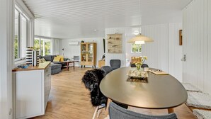 A modern living room with a sofa, dining table, and a dog. There is a cabinet with glass doors and a hanging light fixture. - Cozy home in Ulfborg with kitchen (Ulfborg)