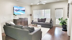 Una sala de estar con un sofá gris, un mueble de madera, un televisor en la pared y un ventilador de techo.