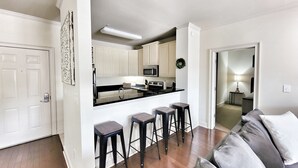 Una cocina moderna con armarios blancos, encimera negra y campana extractora de acero inoxidable. Hay tres taburetes negros y un sofá gris en la zona de estar.