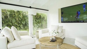 A modern living room with a white sofa, a round coffee table, and a large TV screen displaying a sports game. - Fantastic Golf-View Villa. W/ Jacuzzi, Maid (CASA DE CAMPO)