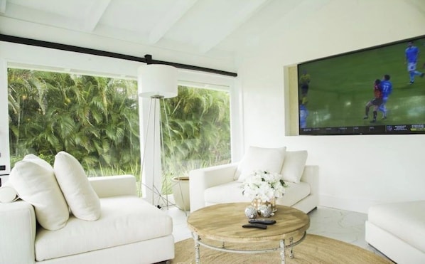 Una sala de estar moderna con un sofá blanco, una mesa de centro redonda y un televisor grande mostrando un partido deportivo.