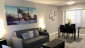 Ein Wohnzimmer mit grauem Sofa, einem gelben Sessel, einem schwarzen Couchtisch und einem Essbereich mit schwarzem Tisch und Stühlen.