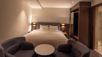 A hotel room with a large bed, bedside tables, a TV, a wardrobe, and two armchairs.