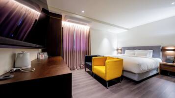 Una habitación de hotel moderna con una cama, un escritorio con cafetera, un sillón amarillo y un televisor en la pared.