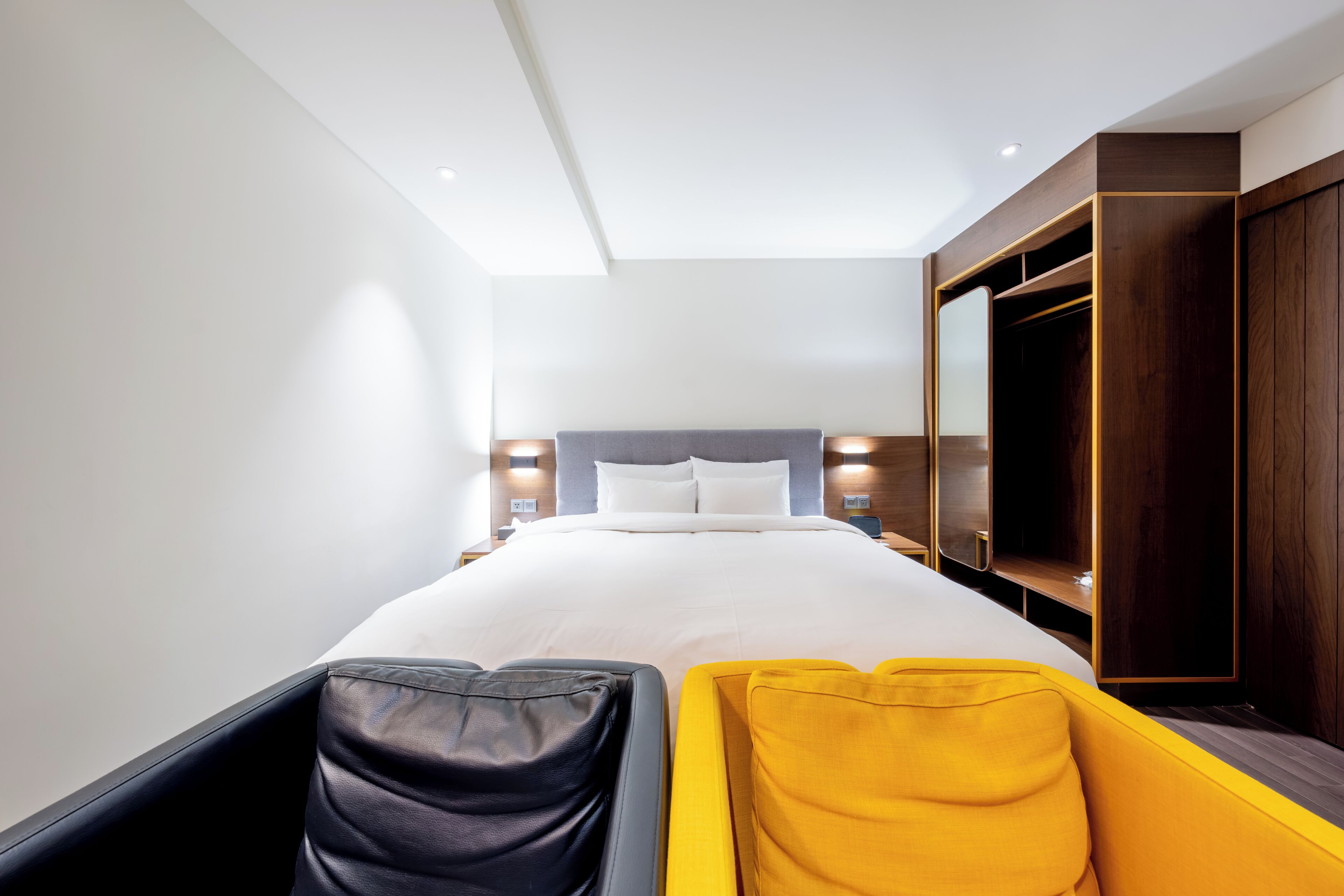 A modern hotel room with a large bed, a wardrobe, and a seating area with a yellow and black chair.