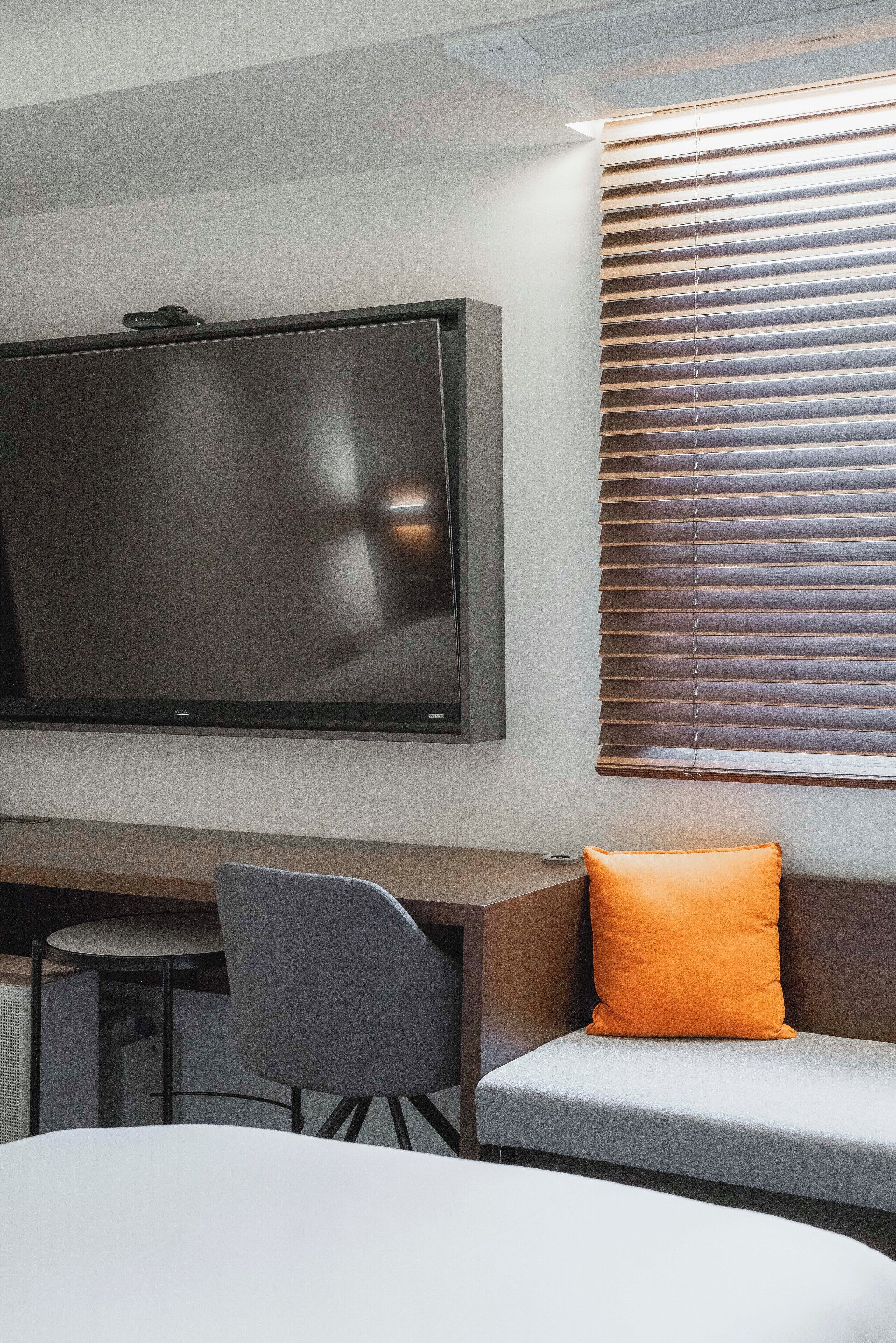 A modern hotel room with a large flat-screen TV mounted on the wall, a wooden desk, a grey chair, and a grey sofa with an orange cushion.