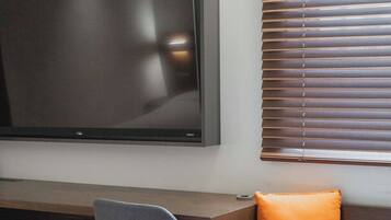 A modern hotel room with a large flat-screen TV mounted on the wall, a wooden desk, a grey chair, and a grey sofa with an orange cushion.