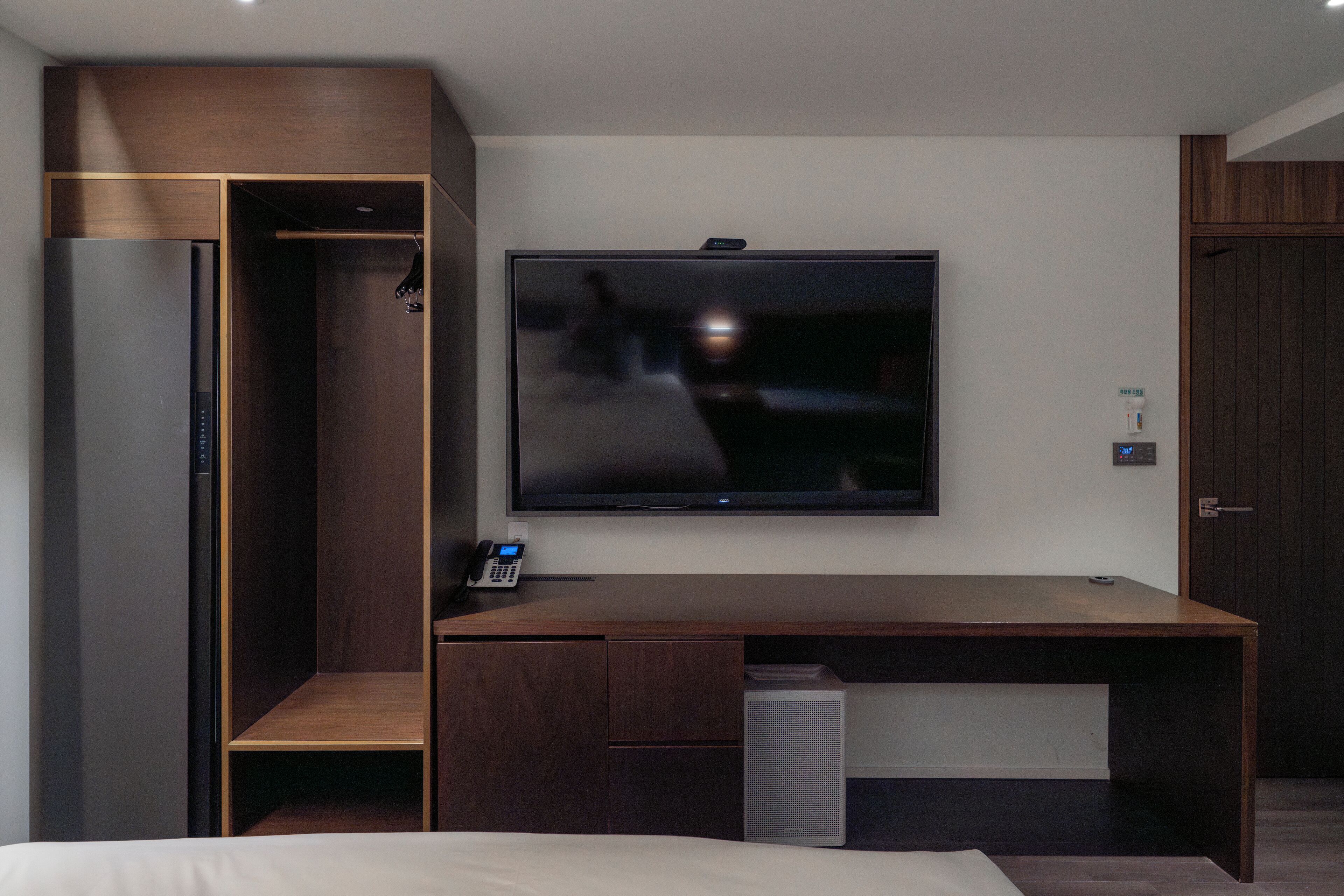 A modern hotel room with a large bed, a wooden desk, a flat-screen TV mounted on the wall, and a built-in wardrobe.