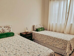 A room with two beds, each with a patterned bedspread, a bedside table with a lamp, and a window with curtains.
