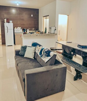 A modern living room with a grey sofa, a dining table, and a kitchen area in the background.