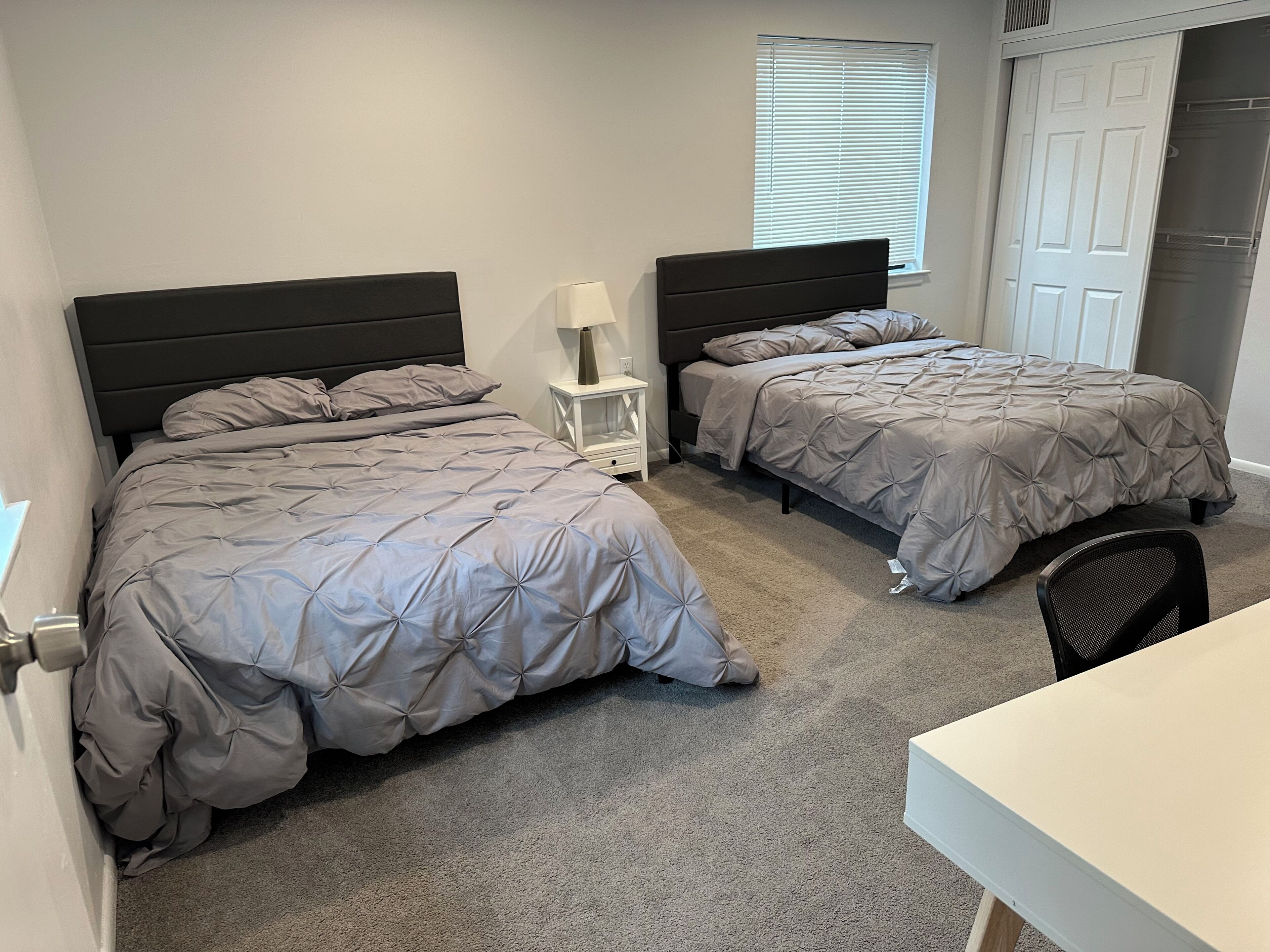 Een slaapkamer met twee bedden, een nachtkastje, een bureau en een raam met jaloezieën.